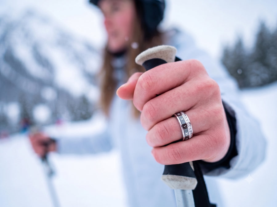 A bare hand gripping a ski pole in deep snow, wearing the Luna ring, a mountain slope behind.