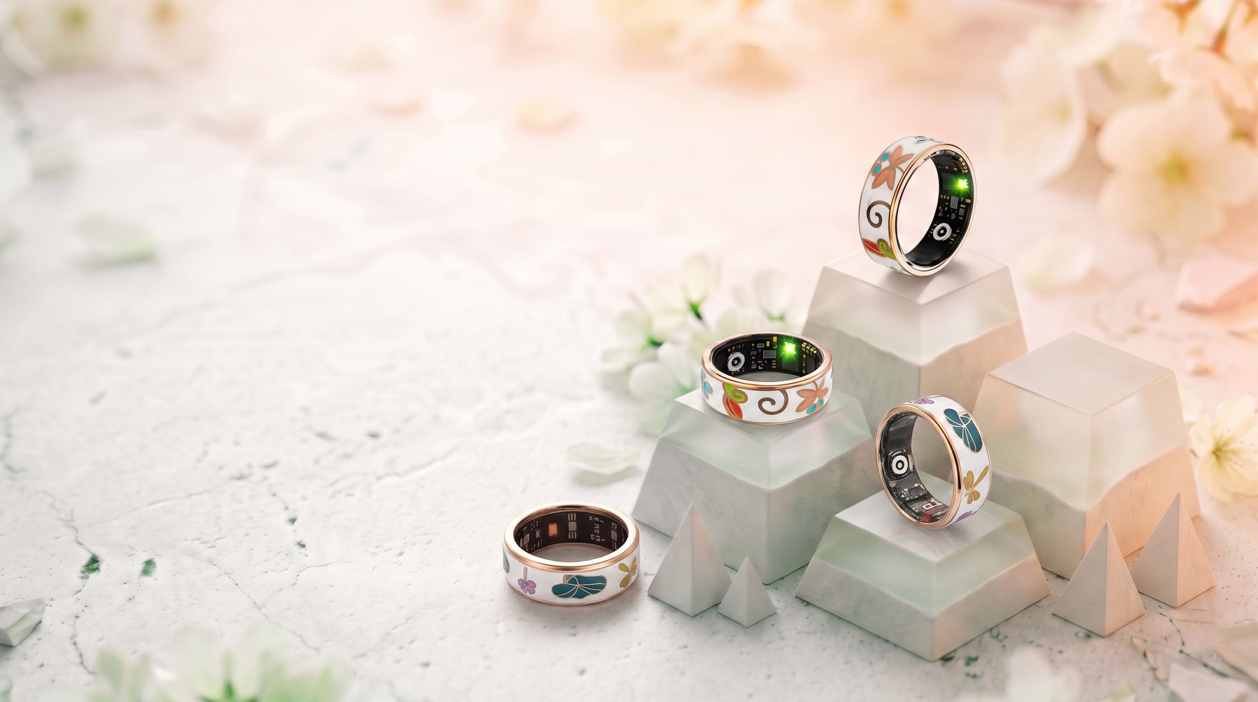 Four Luna smart rings in white enamel with floral dragonfly artwork and rose gold edges, resting on pale stone blocks among white blossom.