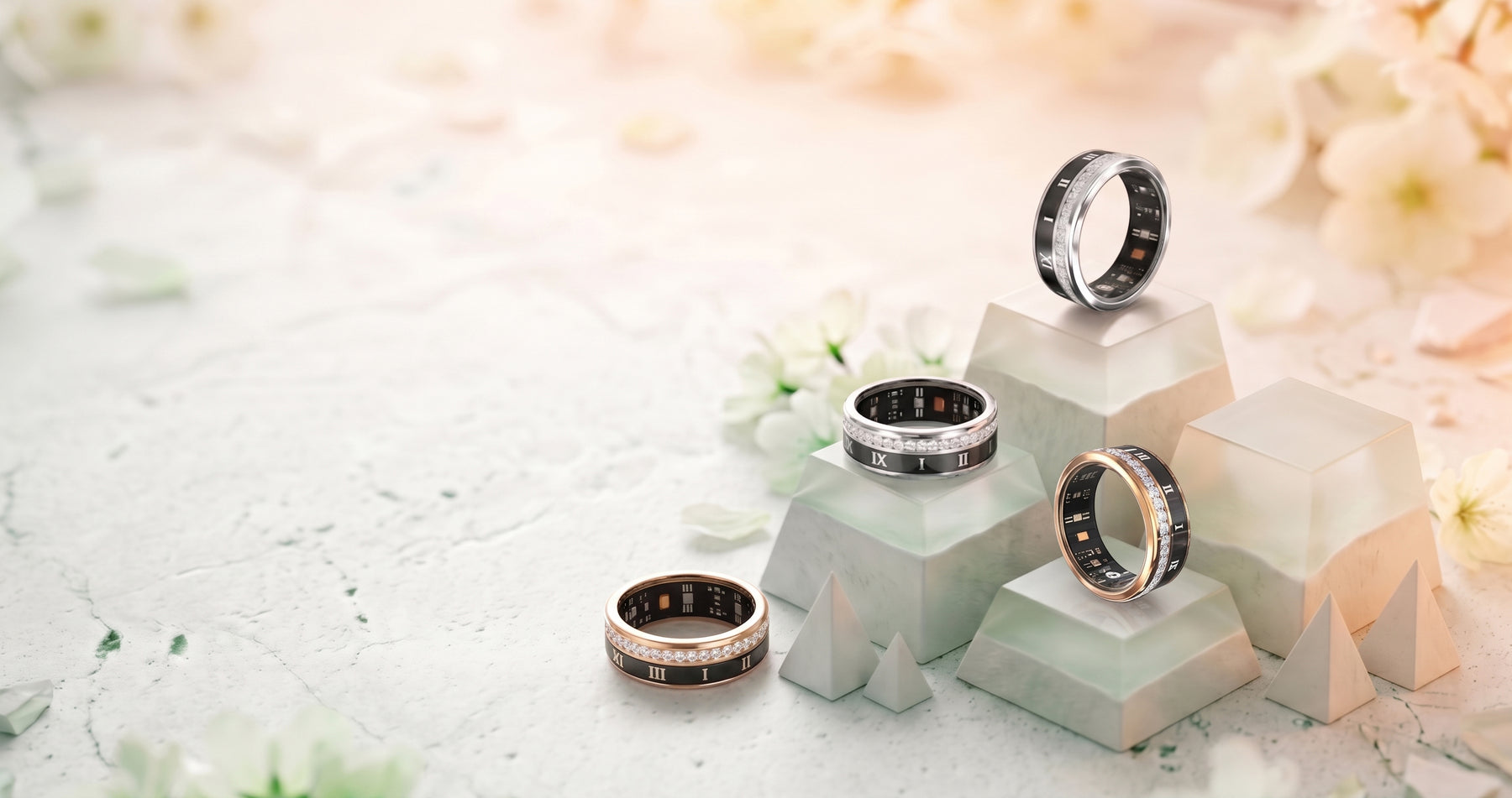 Four Luna smart rings in rose gold and silver with black enamel, resting on pale stone blocks among white blossom.
